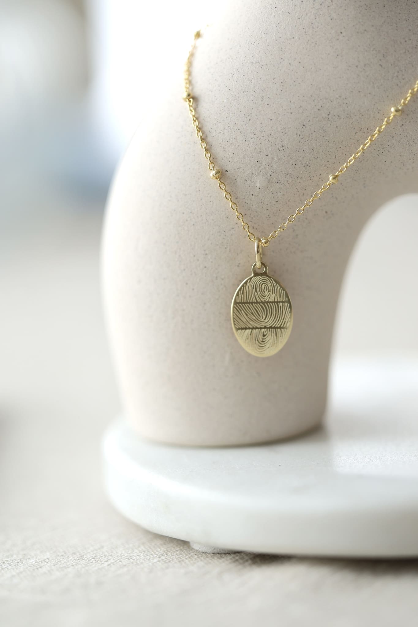 Halskette in Gold mit 3 Fingerabdrücken graviert. Die Kette hat kleine Kugeln und hängt an einer Vase.