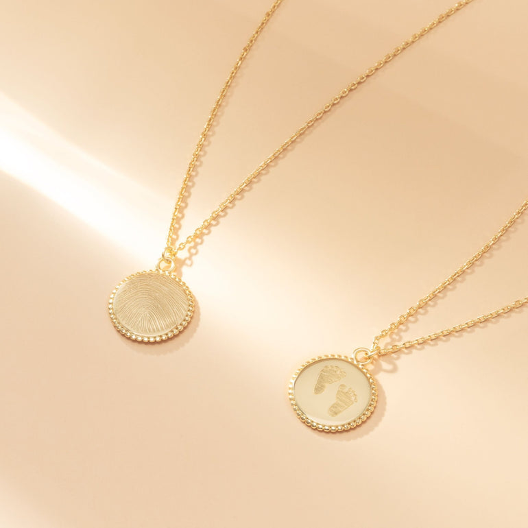 Zwei 18kt vergoldete Vintage Coin Halsketten auf beigen Hintergrund mit Lichteinfall. Auf einer Halskette ist ein Fingerabdruck-, auf der anderen Halskette sind zwei Fußabdrücke graviert.